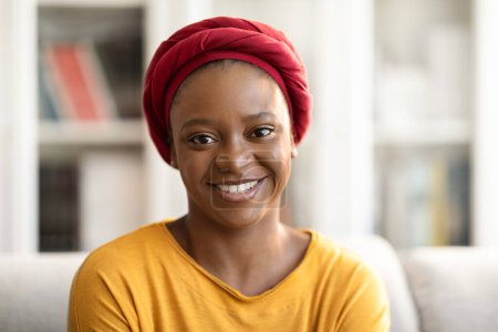 Foto de Portrait of cheerful pretty young black woman wearing red turban posing at home, sitting on couch and smiling at camera, free space. Millennials, women, females lifestyle concept - Imagen libre de derechos