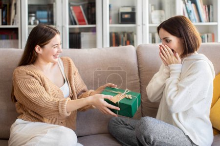 Photo for Glad european millennial female gives gift box to surprised girlfriend, congratulate with Birthday in living room interior. Celebration holiday, Valentine day, anniversary together at home, emotions - Royalty Free Image
