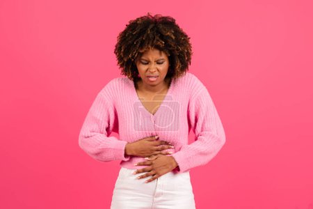 Foto de Sad pretty young black curly woman in casual suffering from stomach hurt, menstrual pain, isolated on pink background. Female health problems, poisoning, indigestion, gastrointestinal diseases, nausea - Imagen libre de derechos