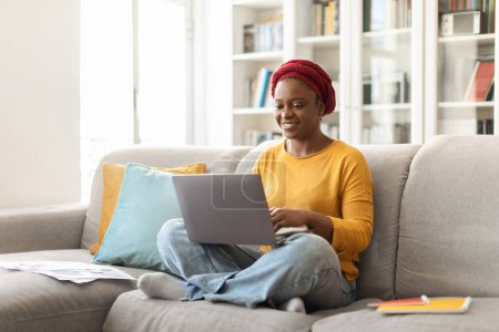 Téléchargez les photos : Positive pretty millennial african american woman with red turban in casual outfit sitting on couch, typing on laptop keyboard and smiling, female digital nomad working from home, copy space - en image libre de droit