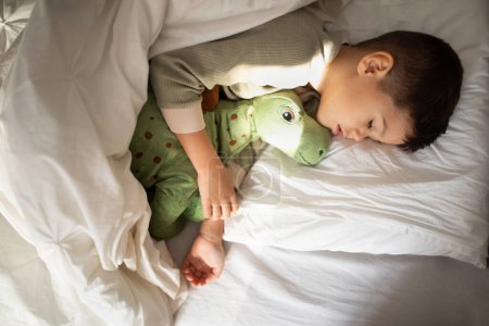 Photo for Happy cute tired little boy lies on white comfort bed with closed eyes, hugs toy dinosaur, sleeping in bedroom interior, top view, free space, close up. Health care, childhood, rest, relax at night - Royalty Free Image