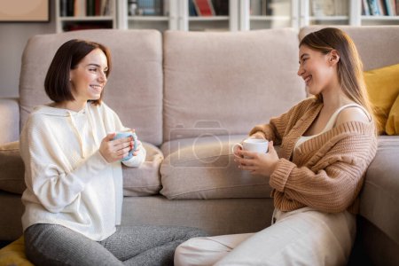 Photo for Laughing european millennial girlfriends in sweaters with coffee cups sit on sofa, enjoy meeting and talking in comfort living room interior. Gossip, good news, communication together, relationships - Royalty Free Image