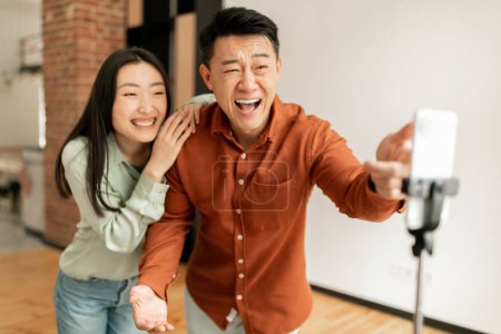 Photo for Overjoyed asian couple bloggers broadcasting from home, sharing their lifestyle with followers, talking and laughing at smartphone camera, living room interior - Royalty Free Image