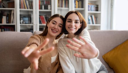 Foto de Cheerful european millennial girlfriends in sweaters make selfie for blog and social media, wave hand and show peace sign in living room interior. Chat, video shooting, stream together, blog at home - Imagen libre de derechos