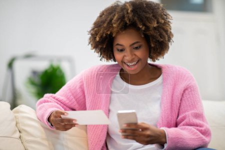 Photo for Giveaway, lottery, online bet. Emotional pretty young african woman in casual outfit resting alone on couch at home, using brand new smartphone, checking her lottery ticket online, copy space - Royalty Free Image