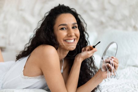 Foto de Cheerful arab millennial curly lady in domestic clothes lies on bed, doing basic makeup with mascara, cosmetics and mirror in bedroom interior. Morning alone at home, beauty care and daily procedures - Imagen libre de derechos