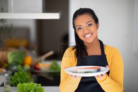 Téléchargez les photos : Cheerful millennial african american lady chef in apron show ready dish of fried fish at table with organic fresh vegetables in white kitchen interior. Household chores, cooking dinner, health care - en image libre de droit