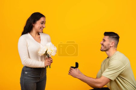 Photo for Cheerful young arab guy in casual on knee gives box with ring to woman with flowers, isolated on yellow background, studio. Love, relationship, proposal, engagement, ad and offer, emotions of bride - Royalty Free Image