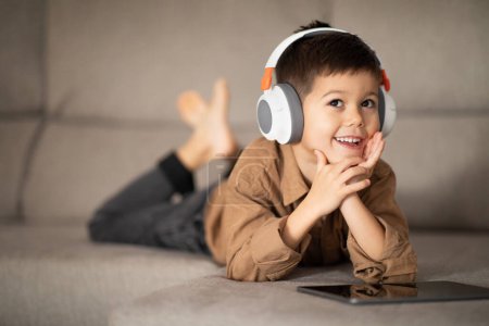 Téléchargez les photos : Happy pensive little boy in wireless headphones thinks, creative idea, watching video on tablet, lies on sofa in room interior. Study, upbringing and childhood, rest with a device and app at home - en image libre de droit