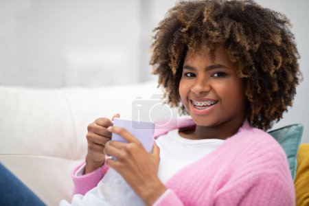 Téléchargez les photos : Happy attractive curly african american young woman with teeth braces in comfy casual outfit chillng alone at home, reclining on couch, drinking coffee and smiling at camera, closeup, copy space - en image libre de droit