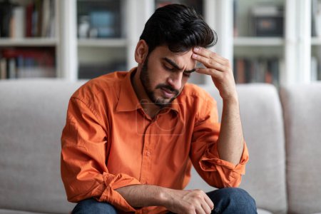 Photo for Upset unhappy frustrated young middle eastern man in smart casual sitting on couch at home, touching his head and looking down, experiencing lifetime crisis, become depressed, copy space - Royalty Free Image