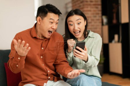Téléchargez les photos : Portrait of overjoyed asian couple using smartphone and making winner sign gesture, shaking fists, sitting on couch at home, reading great news - en image libre de droit