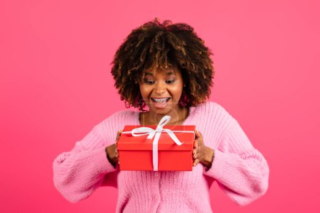 Foto de Cheerful shocked excited young black curly lady in casual with braces looks at box with gift, isolated on pink background, studio. Dreams, surprise, present and congratulations on birthday and holiday - Imagen libre de derechos