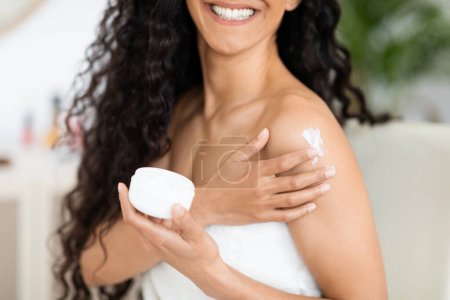 Téléchargez les photos : Cheerful islamic millennial curly lady in towel holding jar with copy space, apply cream on shoulder, enjoy procedure in bedroom interior. Beauty care, spa day, skin moisturizing, treatments at home - en image libre de droit