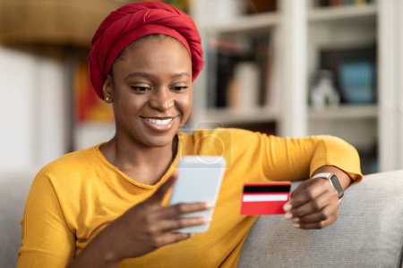 Foto de E-commerce. Cheerful beautiful millennial african american woman in red turban using plastic credit card and cell phone at home, paying for goods and services, shopping online, closeup, copy space - Imagen libre de derechos
