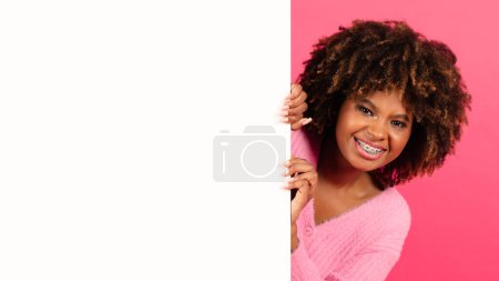 Téléchargez les photos : Smiling pretty young black curly female in casual with braces peeks out from behind banner with empty space isolated on pink background, studio, panorama. Recommendation and advice, sale, ad and offer - en image libre de droit