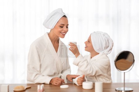 Photo for Mother and daughter in bathrobes doing skin care routine at home, happy beautiful mom and cute preteen female child sitting at beauty table, applying face cream and having good time together - Royalty Free Image