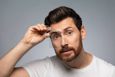 Téléchargez les photos : Eyebrow and hair removal of man for skincare. Middle aged bearded man tweezing eyebrows, standing over grey studio background. Cosmetics and self care - en image libre de droit