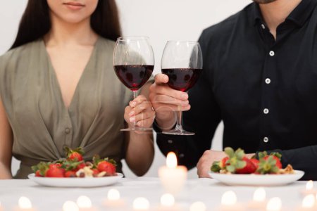 Téléchargez les photos : Cropped of young couple in love man and woman wearing nice festive outwear cheering with glasses of red wine, lovers celebrating anniversary at restaurant, lit candles, plates with strawberry on table - en image libre de droit