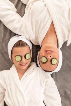 Téléchargez les photos : Spa Day. Happy Little Girl And Her Mother Wearing Bathrobes Lying With Cucumber Slices On Eyes, Cheerful Family Mom And Daughter Doing Skincare Treatments And Having Fun Together At Home, Top View - en image libre de droit