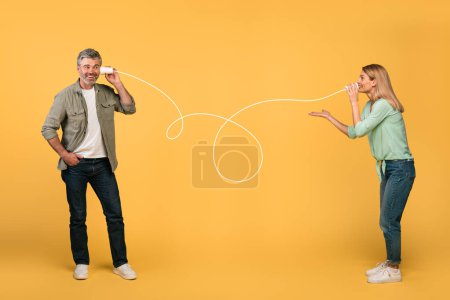 Téléchargez les photos : Communication and fun. Happy middle aged spouses talking through tin can telephone, standing over yellow studio background. Man and woman hearing and speaking through cups - en image libre de droit