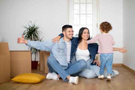 Happy young family with little son having fun at new home after moving, smiling millennial parents playing with their child while sitting on floor among cardboard boxes, opening arms for embrace
