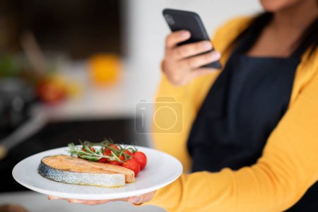 Téléchargez les photos : Young african american lady chef in apron makes photo of fresh fish on phone in kitchen interior, blurred, cropped. Professional cooks at home, healthy eat, diet, food blog and app for social networks - en image libre de droit