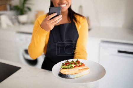 Photo for Cheerful millennial african american lady in apron taking photo of dish of fish on smartphone in white kitchen interior, cropped. Cooking dinner, health care and food blog, app for professional work - Royalty Free Image