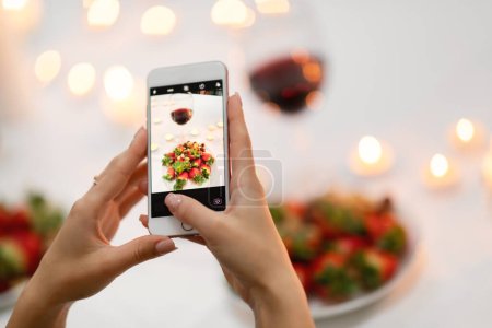Téléchargez les photos : Cropped of lady taking photo of glass with red wine and plate full of strawberry on festive table decorated with lit candles, woman sharing photos on social media while romantic dinner, using phone - en image libre de droit