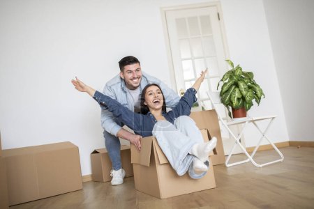 Photo for Happy Young Couple Having Fun Together While Moving To New Home, Joyful Millennial Woman Riding In Cardboard Box In Living Room Interior, Loving Family Fooling In Their Apartment After Relocation - Royalty Free Image