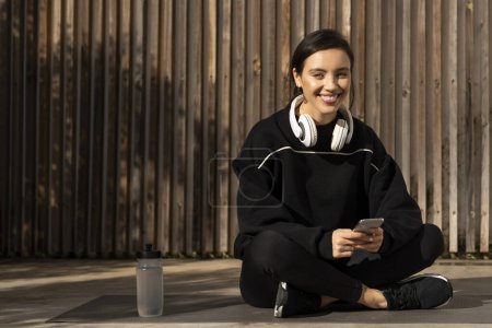 Téléchargez les photos : Happy young european woman athlete in sportswear and headphones sits on floor with smartphone, enjoys weight loss, fitness in city, outside. Breack, relax from muscle training, body and health care - en image libre de droit