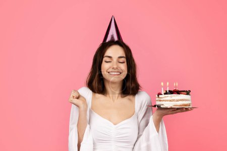 Foto de Cheerful young caucasian lady with closed eyes in hat hold cake with candles and dreaming, enjoy feast isolated on pink background, studio. Emotions from celebrating holiday, make wish for birthday - Imagen libre de derechos