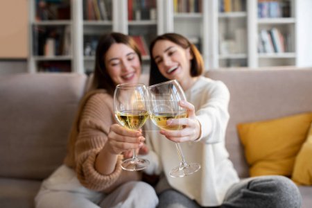 Foto de Cheerful pretty european millennial women in sweaters cheers glasses of wine, enjoy relax and free time, celebrating holiday in comfort living room interior. Toast, date and party together at home - Imagen libre de derechos