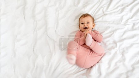 Téléchargez les photos : Cute little baby girl having fun and smiling, lying on bed at home, panorama with free space, top view. Adorable infant kid playing with her toes and laughing - en image libre de droit