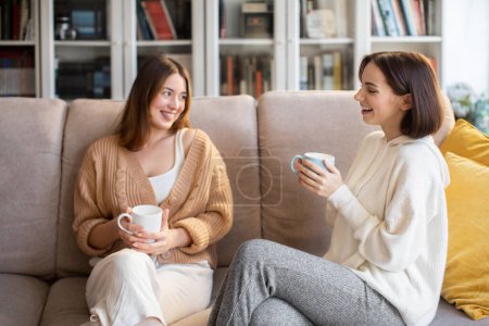 Photo for Laughing european millennial ladies in sweaters with coffee cups talk, sit on sofa, enjoy visit, free time in comfort living room interior. Gossip, communication together, relationships and friendship - Royalty Free Image