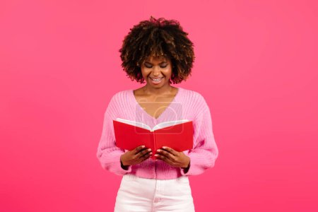 Téléchargez les photos : Cheerful smart pretty young black curly female student in casual reading book, diary, plans day with notepad isolated on pink background, studio, copy space. Study, knowledge, education and literature - en image libre de droit