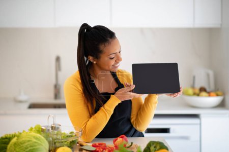 Photo for Cheerful millennial african american female in apron show tablet with empty screen at table with organic fresh vegetables in white kitchen interior. Household chores, cook dinner with app, food blog - Royalty Free Image