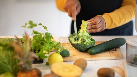 Téléchargez les photos : Millennial african american lady in apron cuts broccoli at table with fresh vegetables in white kitchen interior, close up, cropped. Prepare food, professional shef work, cook dinner, household chores - en image libre de droit