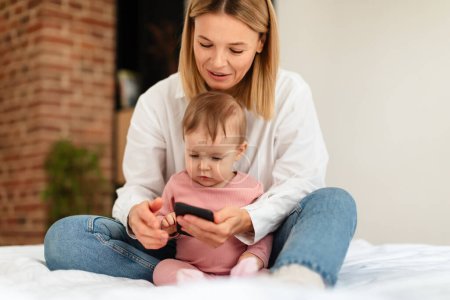 Foto de Happy mom and her little daughter using smartphone, sitting on bed at home, browsing internet or watching videos online while spending time together - Imagen libre de derechos