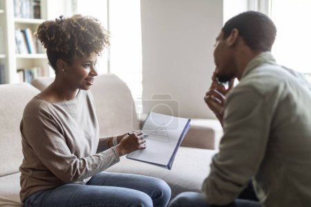Photo for Attractive young black woman psychologist having conversation with male patient, showing him notes in medical chart, explaining mental processes. Depressed guy attending therapy session, copy space - Royalty Free Image