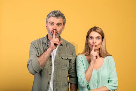 Téléchargez les photos : Keep silence concept. Middle aged spouses gesturing hush sign and looking at camera, standing over yellow studio background. Couple holding finger on lips, silent gesture, secret - en image libre de droit