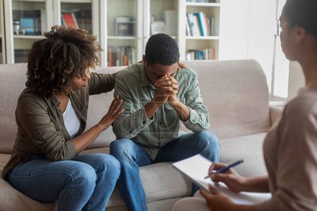 Téléchargez les photos : Loving attentive young african american woman hugging comforting her depressed spouse or boyfriend suffering from anxiety, black couple having family therapy with woman psychologist, clinic interior - en image libre de droit