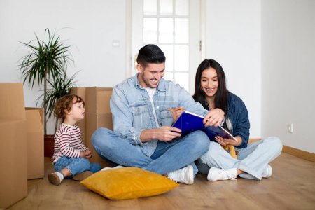 Photo for Happy Young Family With Little Son Making List Of Necessities After Moving To New Home, Cheerful Parent Writing In Notebook While Sitting With Their Kid On Floor Among Cardboard Boxes, Free Space - Royalty Free Image