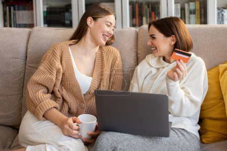 Photo for Laughing happy european millennial ladies shopaholic in sweaters with credit card, laptop shopping online in comfort living room interior, copy space. Good sale, cashback and finance banking at home - Royalty Free Image
