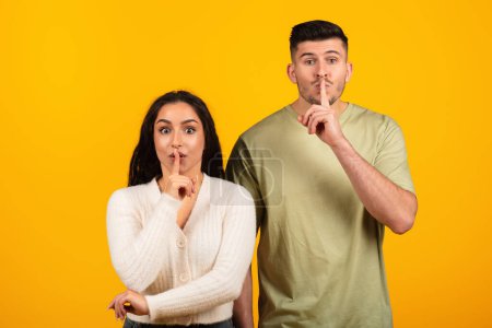 Photo for Cheerful inspired pretty young arabic woman and guy in casual put finger to lips, make shhh sign, isolated on yellow background, studio. Secret gesture, gossip emotions, good news, sale, ad and offer - Royalty Free Image