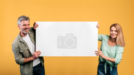 Téléchargez les photos : Cheerful middle aged couple holding empty board for advertisement or text and smiling, yellow studio background. Happy man and woman with blank placard for advert - en image libre de droit