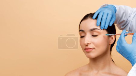 Foto de Cosmetologist Doctor Making Botox Injection For Eyes Area To Young Indian Woman, Attractive Hindu Lady Getting Anti-Wrinke Treatment While Standing Over Beige Studio Background, Copy Space - Imagen libre de derechos