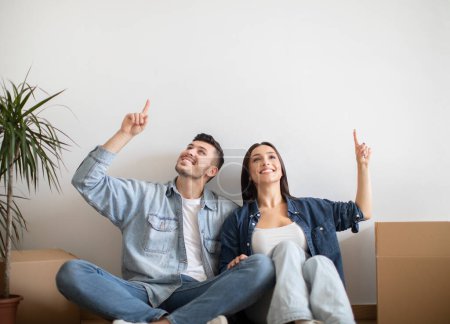 Foto de Happy Young Spouses Sitting On Floor After Moving And Pointing Up, Smiling Millennial Couple Showing Free Copy Space Above Their Heads, Dreaming About Interior Design In New Home, Copy Space - Imagen libre de derechos