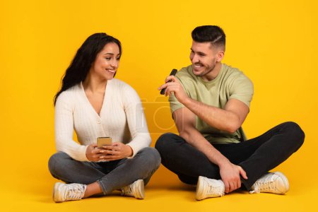 Téléchargez les photos : Cheerful young arab male in casual shows phone to wife, sit on floor isolated on yellow background, studio. Recommendation app for blog and social networks, sale, love, relationship, ad and offer - en image libre de droit
