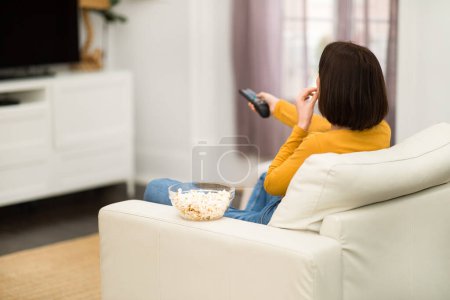 Téléchargez les photos : Rear view of brunette young woman sitting on couch in cozy living room, watching TV show, movies with popcorn, holding remote controller, spending time alone at home, copy space - en image libre de droit
